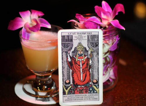 A cocktail with a pink hue beside a tarot card featuring a figure in red, adorned with orchids in a jar.