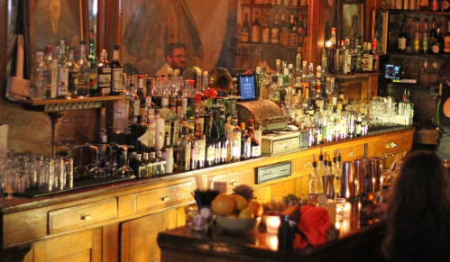 A dimly lit bar with a wide array of bottles on the shelves and a wooden counter, creating a cozy atmosphere.