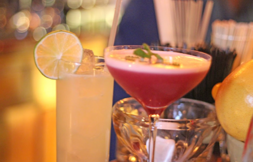 Two colorful cocktails on a bar: a yellow drink with a lime slice and a pink drink garnished with mint.