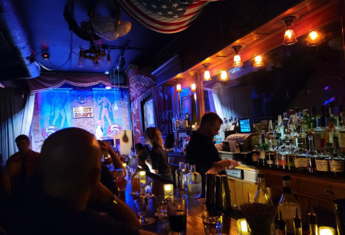 A dimly lit bar scene with patrons, a well-stocked bar, and colorful lights creating a lively atmosphere.