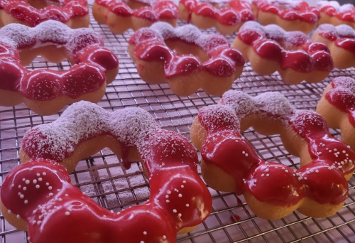 A tray of ring-shaped donuts topped with red icing and powdered sugar, cooling on a wire rack.