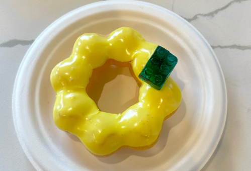 A yellow frosted donut shaped like a flower, topped with a green Lego piece, on a white paper plate.