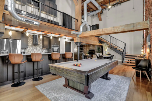 A stylish loft with a pool table, modern kitchen, and wooden beams, featuring a staircase and cozy seating area.