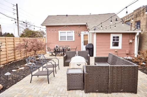 A cozy backyard with a patio, seating area, grill, and string lights, surrounded by a wooden fence and landscaping.