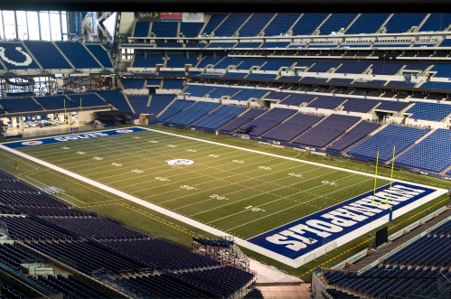 A wide view of an empty football stadium with a green field and blue seating, featuring the "Indianapolis" logo.