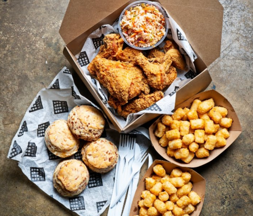 A spread of fried chicken, coleslaw, tater tots, and biscuits in takeout boxes on a rustic surface.