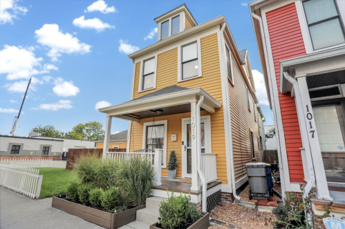 A three-story yellow house with a porch, surrounded by greenery and neighboring red and white houses.