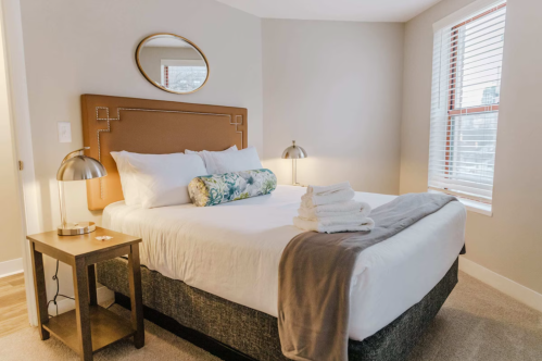 Cozy bedroom featuring a large bed, decorative pillows, bedside lamps, and neatly stacked towels on a side table.