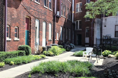 A sunny courtyard with greenery, a chair, and bicycles near brick buildings.