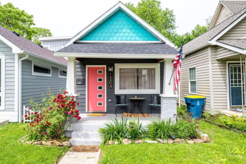 A charming house with a teal roof, pink door, and a small porch surrounded by greenery and flowers.