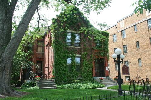 A brick house covered in green ivy, surrounded by trees and a well-kept garden, with a nearby brick building.