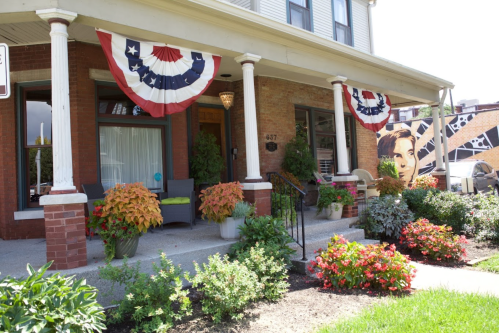 A charming house with patriotic bunting, colorful flowers, and a mural on the side, set in a sunny neighborhood.