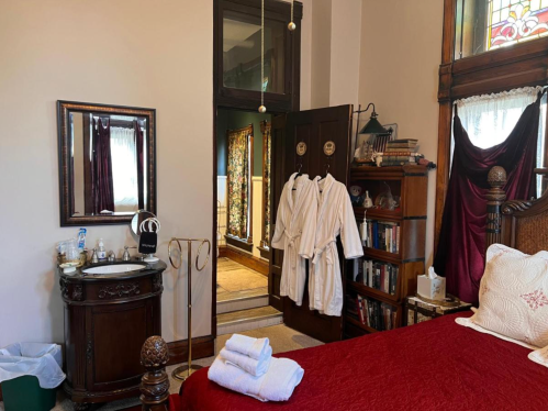 Cozy bedroom with a red bedspread, vintage furniture, and robes hanging by the door, leading to a bright hallway.
