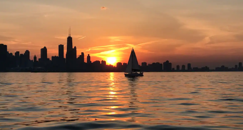 A sailboat glides across a calm lake at sunset, with a city skyline silhouetted against the colorful sky.