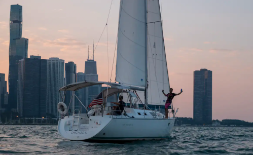 A sailboat with a person waving, set against a city skyline at sunset.