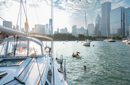 A sailboat in the foreground with swimmers in the water, city skyline and boats in the background under a cloudy sky.