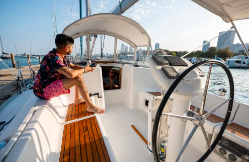 A man in a floral shirt and pink shorts sits on a sailboat, enjoying the view of the city and water.