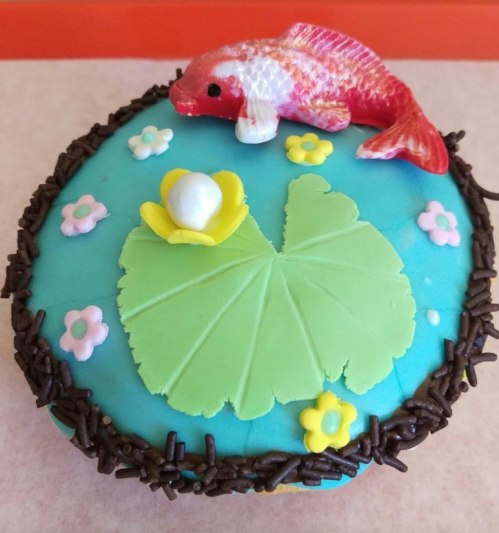 A round cake decorated with blue icing, a green lily pad, a goldfish, and colorful flowers.