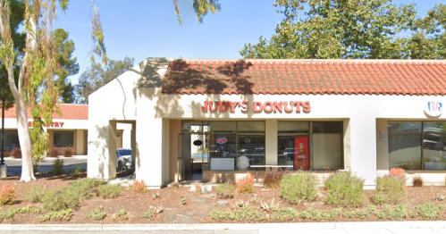 Exterior of Judy's Donuts shop with a red sign, surrounded by greenery and other buildings in a shopping area.