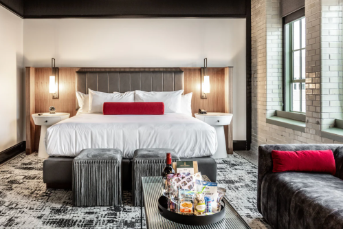 A modern hotel room featuring a king-sized bed, stylish decor, and a seating area with snacks and drinks on a table.