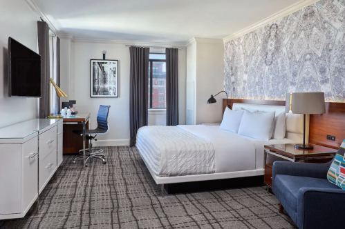 A modern hotel room featuring a queen bed, desk, chair, and stylish decor with large windows and a patterned wall.