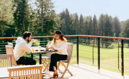 A couple enjoys a meal on a sunny balcony overlooking a green landscape, dressed casually in stylish outfits.
