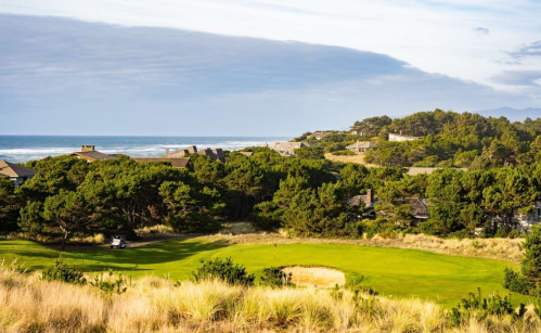 A scenic view of a golf course near the ocean, surrounded by trees and coastal hills under a cloudy sky.