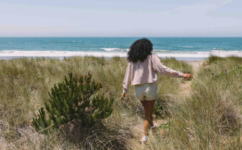 A person walks along a sandy path through tall grass, heading towards the ocean on a sunny day.