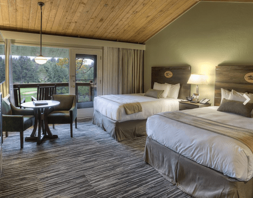 Cozy hotel room with two beds, a table, and a balcony overlooking a scenic view. Warm wood accents and soft lighting.