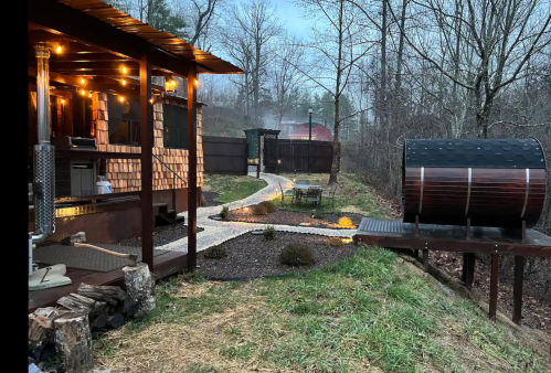 A cozy outdoor space with a wooden cabin, pathway, and a hot tub surrounded by trees in a misty setting.
