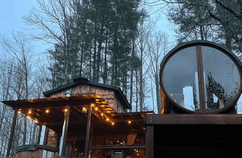 A cozy wooden cabin with a circular window, surrounded by tall trees and soft evening lighting.