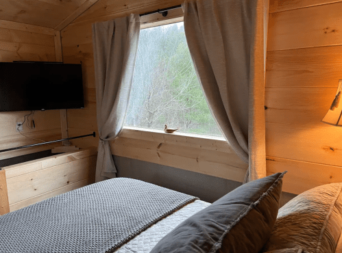 Cozy cabin interior with a bed, window with curtains, and a TV on the wall, surrounded by wooden walls.