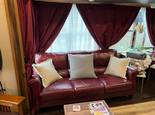 A burgundy leather couch with three pillows, set against a window with sheer curtains and a cozy interior.