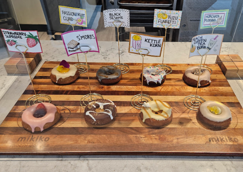A display of colorful donuts on a wooden board, each labeled with unique flavors like strawberry, s'mores, and horchata.
