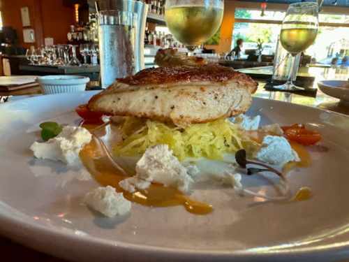 A plated dish featuring grilled fish atop spaghetti squash, garnished with cheese, cherry tomatoes, and a sauce.