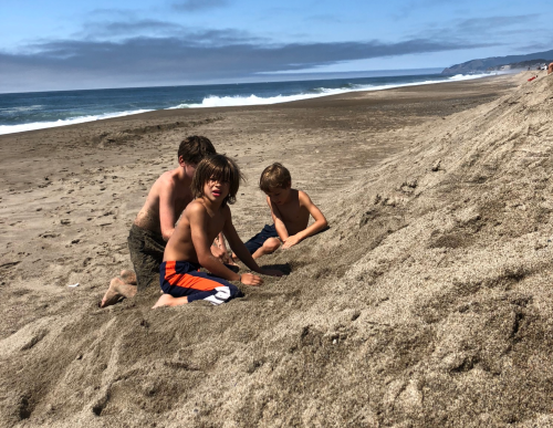 Three children playing in the sand on a beach, with waves and a cloudy sky in the background.