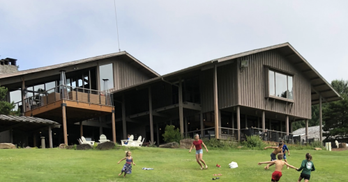 Children running on a grassy hill in front of a large wooden building with a deck and trees in the background.