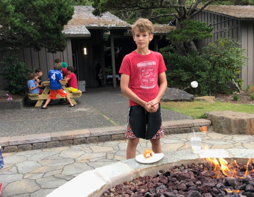 A boy stands by a fire pit holding a plate of food, with children playing in the background at a camp setting.