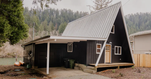 A black A-frame house with a metal roof, surrounded by trees and gravel driveway, near a river.