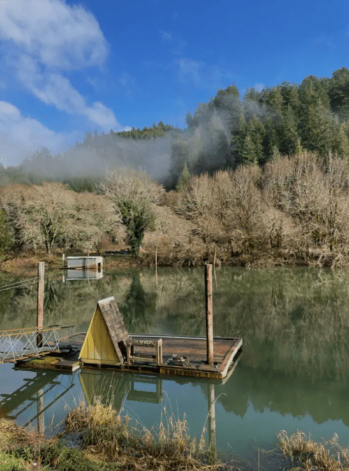 A serene lake scene with a small dock, surrounded by trees and misty hills under a blue sky.