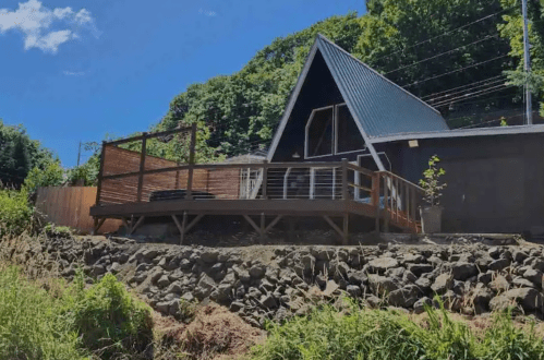 A modern A-frame house with a wooden deck, surrounded by greenery and blue skies.