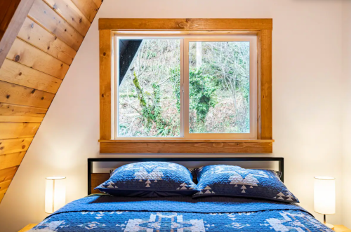 Cozy bedroom with a blue patterned bedspread, two lamps, and a large window overlooking greenery.