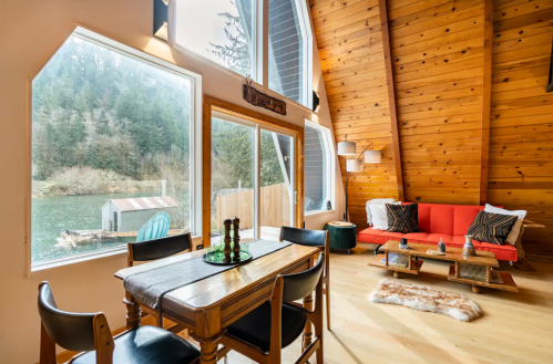 Cozy interior with wooden walls, a dining table, and a sofa, overlooking a serene lake through large windows.