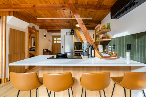 Modern kitchen with wooden beams, a white island, brown bar stools, and green tile accents. Cozy and stylish design.