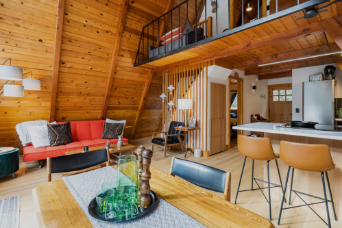 Cozy A-frame interior with wooden walls, a red sofa, dining table, and a lofted sleeping area. Modern and inviting design.