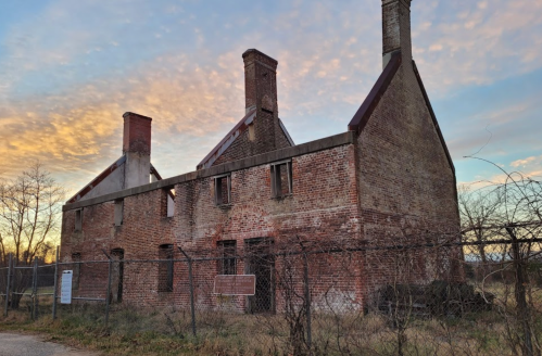 A weathered brick building with two chimneys, surrounded by a fence, against a colorful sunset sky.