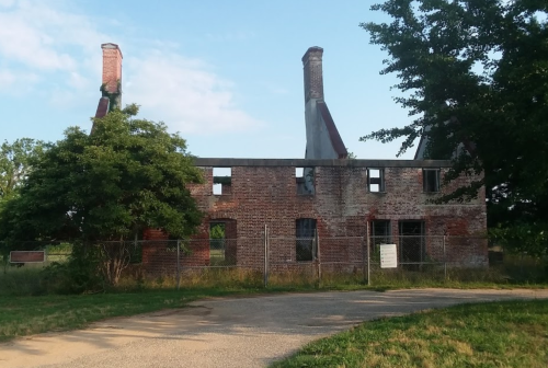 Ruins of a brick building with two tall chimneys, surrounded by a fence and greenery under a blue sky.