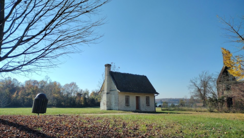 A small, white cottage sits in a grassy area with trees and a clear blue sky in the background.