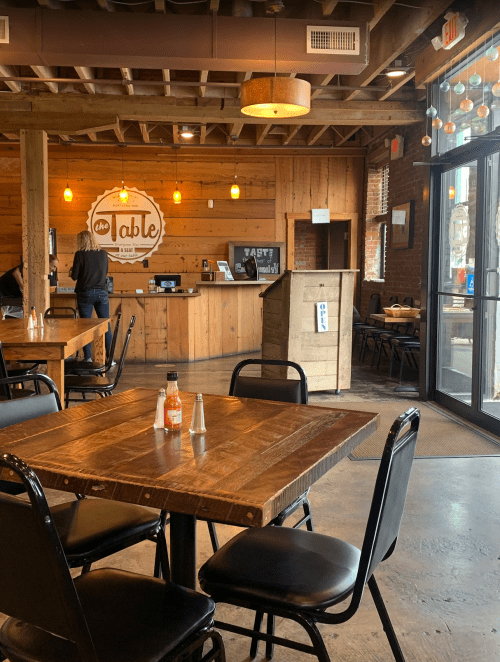 A cozy restaurant interior with wooden walls, tables, and a counter. A person stands at the register.