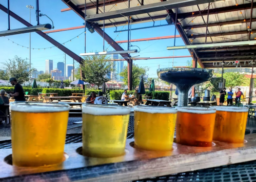 A flight of five craft beers on a wooden paddle, with a city skyline in the background and outdoor seating.